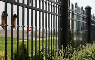 Black metal fence with vertical bars and decorative elements, bordering a grassy area with plants, and a building in the background.