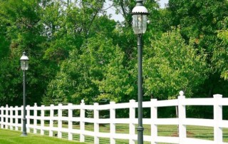 A white wooden fence runs alongside a row of black lampposts on a grassy lawn with dense green trees in the background.