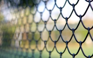 Close-up of a metal chain-link fence with a blurred outdoor background, suggesting a field or park area beyond the fence.