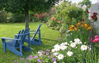 Two blue Adirondack chairs sit on a lawn facing a flower garden with various colorful blooms next to a white picket fence under a leafy tree.