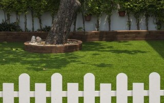 A white picket fence encloses a yard with artificial grass, a large tree in a circular stone bed, and a background of plants growing along a wall.
