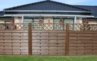 A wooden privacy fence with a lattice top section stands in front of a modern brick house with a dark tiled roof; potted plants hang on the fence.