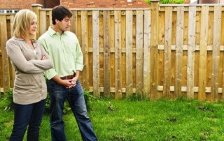 A woman and a man stand on grass in a backyard, looking at the ground near a wooden fence.