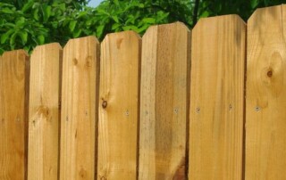 Close-up of a wooden picket fence with vertical slats, set against a background of green leafy trees and a partly cloudy sky.