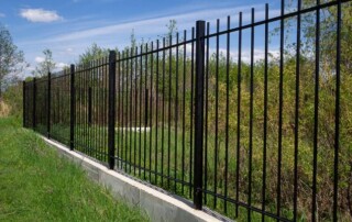 A black metal fence with vertical bars stands on a concrete base, enclosing a grassy area with trees and bushes under a partly cloudy sky.