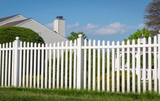 A white picket fence stands in front of a suburban house with shrubs and trees in the background under a clear blue sky.
