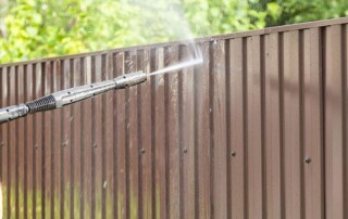 A person uses a pressure washer to clean a brown metal fence outdoors, with green foliage visible in the background.