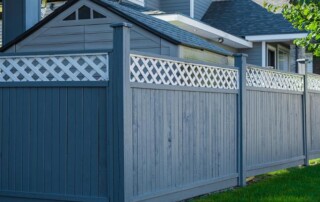 A tall wooden fence with blue-gray panels and white lattice trim runs alongside a house and a small shed, with grass in the foreground.
