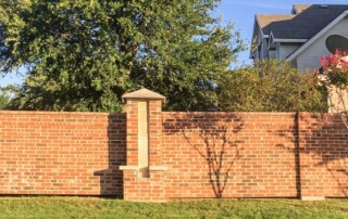 Red brick wall with a decorative pillar in front of a house, trees, and green grass under a clear sky.