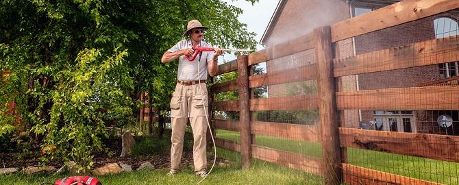 A man wearing a hat and sunglasses uses a pressure washer to clean a wooden fence in a residential yard.