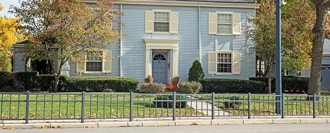 Two-story light blue house with white trim, shutters, and a blue front door. There is a green lawn, trees, shrubs, and a metal fence along the sidewalk.