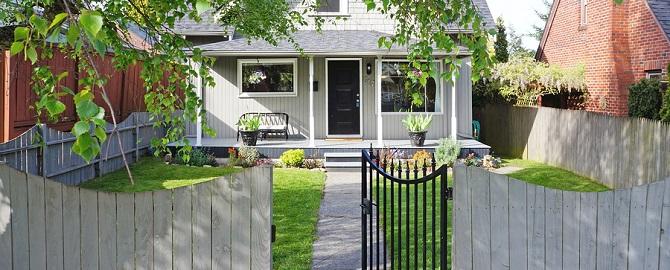 A small house with a front porch, black front door, and fenced yard with green grass, plants, and a garden bench.
