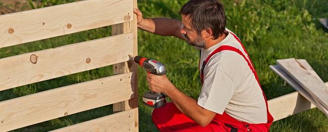 A person in red overalls is using a power drill to assemble a wooden fence outdoors on grass.