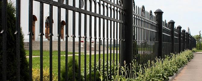Black metal fence with vertical bars runs alongside a sidewalk, with green grass and plants growing at its base.